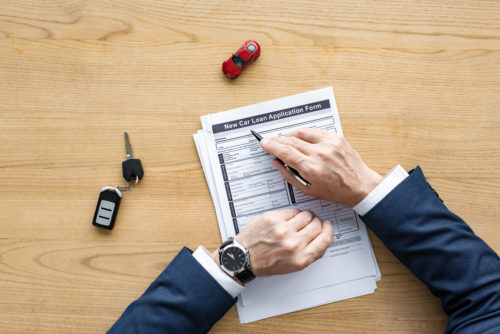 A salesman pointing to the top of a car loan application form with a pen while a set of keys and a model car sit on the desk.