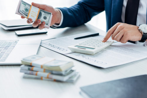 A banker holds a stack of cash, with more on his desk, calculating figures from a contract.