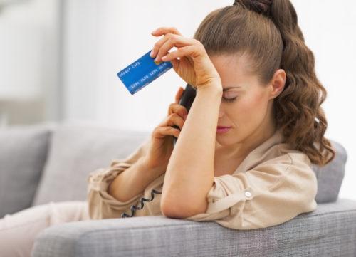 A woman in credit card debt looking somber while talking on the phone and holding her credit card.