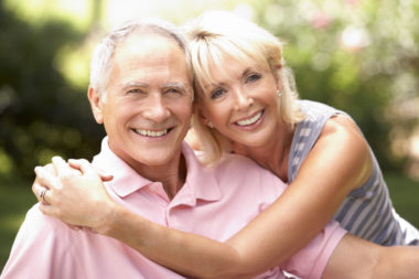 A retired couple hugging in the park while smiling at the camera.