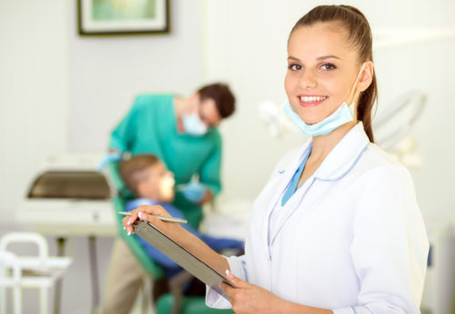 A dental assistant smiles at the camera and holds a clipboard while her colleague works in the background.