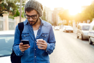 A young man with coffee in one hand and his smartphone open to an investing app in the other.