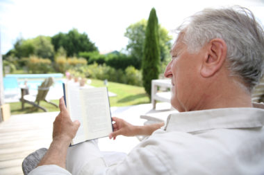 A retired man reading a book in a pool deck chair.