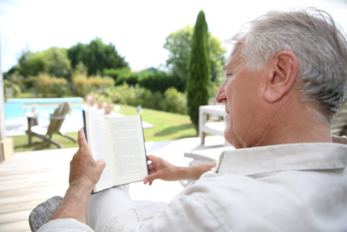 A retired man reading a book in a pool deck chair.