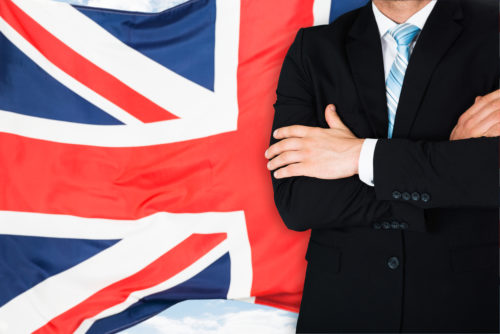 A business,am standing with his arms crossed, in front of the British flag.