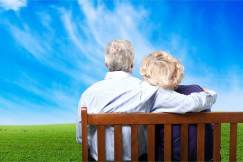 A retired couple on a park bench, looking at the sky over a grassy field.