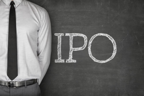 A business man stands next to a chalkboard with the letters "IPO (initial public offering)" written on it.