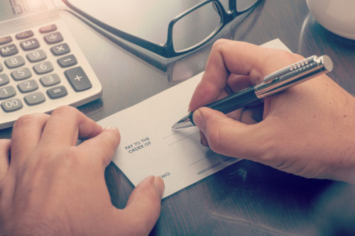 A man writing a check on his desk, which has a calculator and eyeglasses on it.