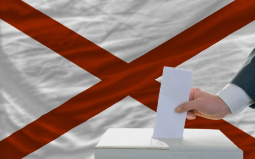 A man putting a ballot in a box with the Alabama state flag in the background.