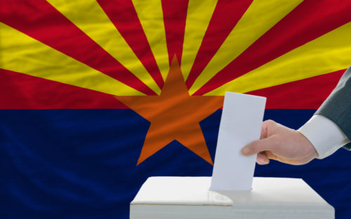 A man putting a ballot in a box with the Arizona state flag in the background.