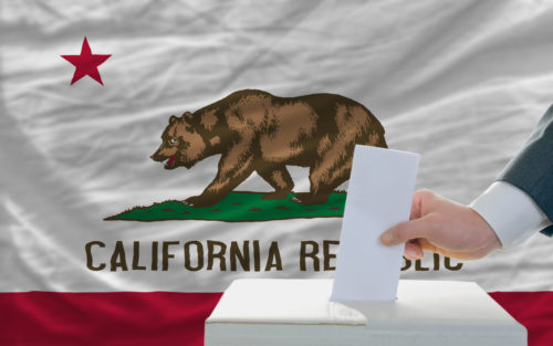 A man putting a ballot in a box with the California state flag in the background.