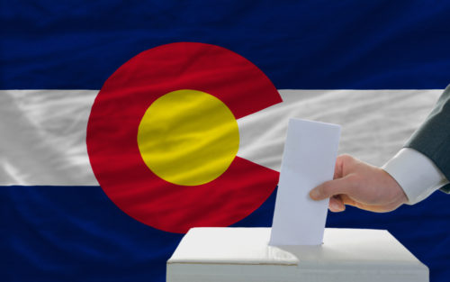 A man puts a vote in a ballet box with the Colorado state flag in the background.