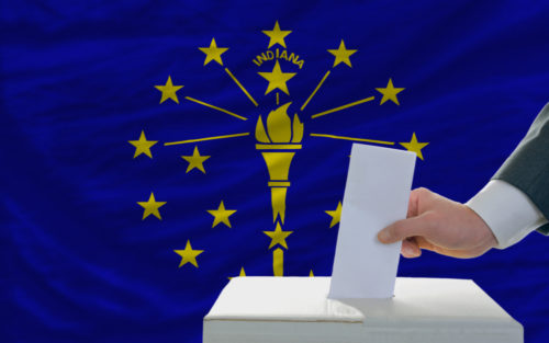 A man putting a ballot in a box with the Indiana state flag in the background.