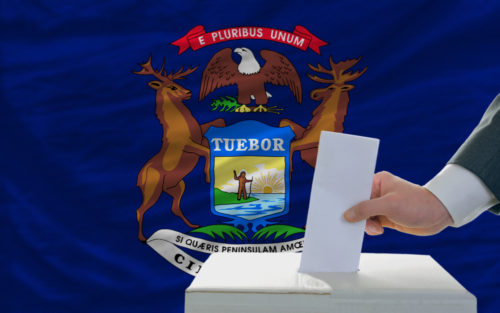 A man putting a ballot in a box with the Michigan state flag in the background.