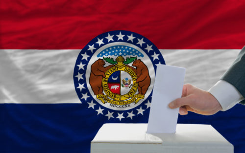 A man placing his vote in a ballot box with the Missouri state flag in the background.