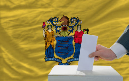 A man putting a ballot in a voting box with the New Jersey state flag in the background.