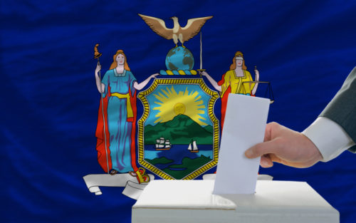 A man placing a ballot in a box with the New York state flag in the background.