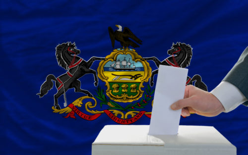 A man putting a ballot in a box with the Pennsylvania state flag in the background.