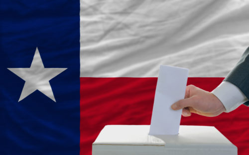 A man places his vote in a ballot box with the Texas state flag in the background.