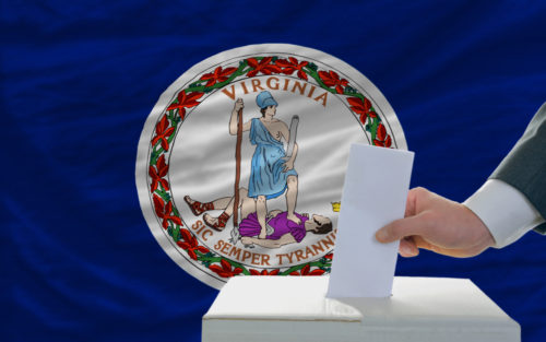 A man putting a ballot in a box in front of the Virginia state flag.