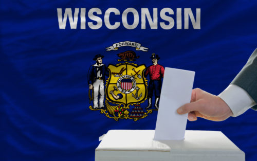 A man places his vote in a ballot box with the Wisconsin state flag in the background.
