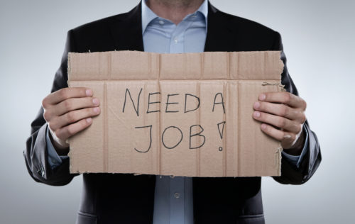 A man in a business suit holds up a cardboard sign that reads "need a job."