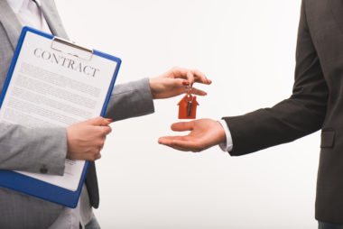 A real estate agent handing a person a set of house keys while holding a clipboard with a document labeled "contract" attached to it.