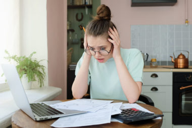 A young woman looks worried calculating her student loans via her laptop and a calculator.
