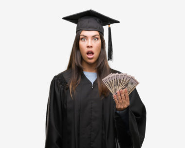 A young woman wearing a graduate uniform looks shocked while holding money.