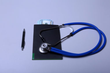 A folder with money sticking out of it lies next to a pen and under a stethoscope.