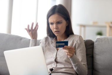 A woman looking upset while on her laptop, holding a credit card in her hand.