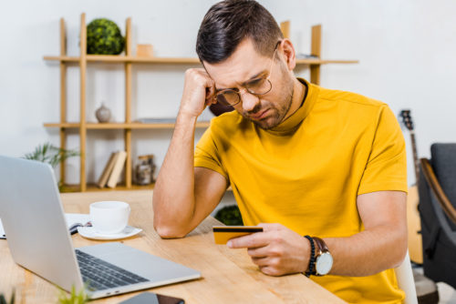 An upset man sitting at his laptop looking at his credit card.