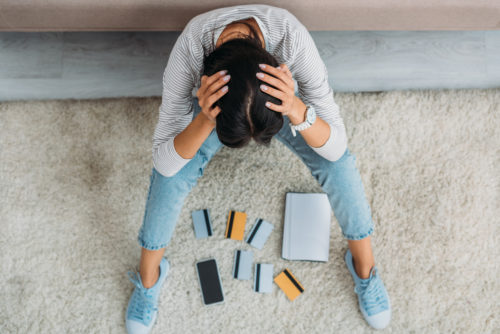 A sad woman sitting on a couch with her head in her hands while credit cards are spread across the floor.