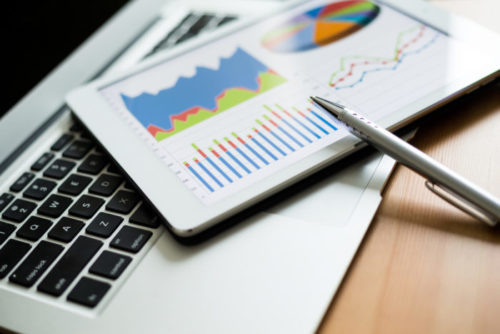 A pen and tablet sit on top of a laptop's keyboard, with the tablet's screen displaying investment figures and charts.