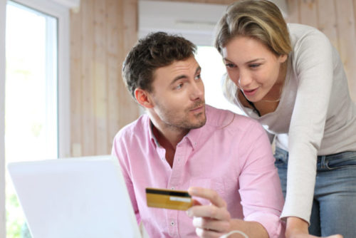 A man sitting at a laptop holding up his credit card for his partner, who is leaning over his shoulder.