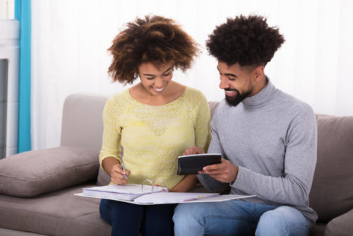 A smiling young couple sitting on a sofa calculating bills to see if they can retire.