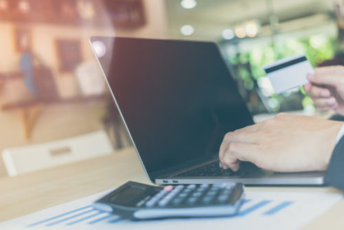 A close up of a man's hands typing on a laptop with a calculator and financial papers on the desk next to him.