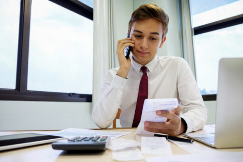 A man speaking on the phone while looking at a receipt at his desk, which has papers, a calculator, and a laptop on it.