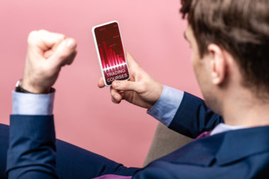 A businessman sits in a pink room while trading stocks on his phone, with a fist raised in the air in excitement.
