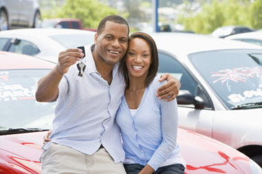 A couple who has just bought a car sit on the hood of if while the man holds out the keys.