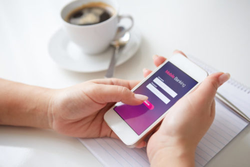 A woman sitting down to coffee while checking her banking account on her phone.