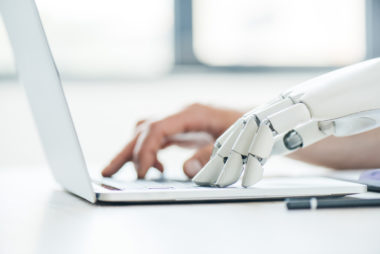 A robot hand and a human hand typing on a laptop.