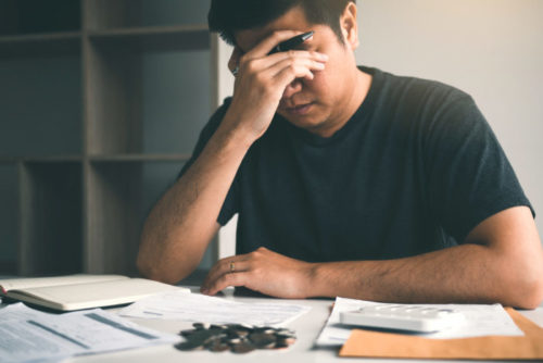 A stressed man puts his head in his hand while invoices clutter the table he is sitting at.