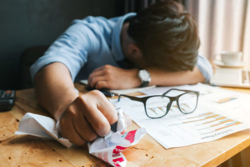 A man has taken off his glasses and set them upon some financial papers while he slumps his head in his arm and crumples another financial document.