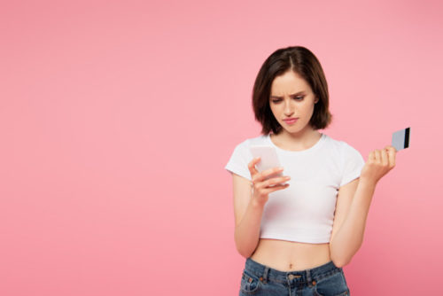 A woman looks disappointed as she checks her smartphone and holds her credit card.