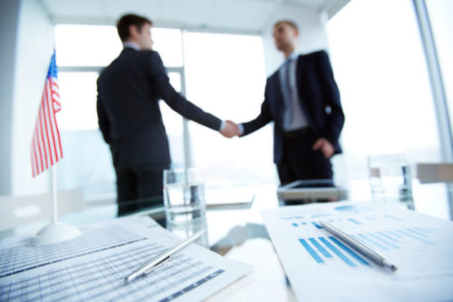 A banker shakes a client's hand in the background while financial papers and an American flag is on his desk in the foreground.