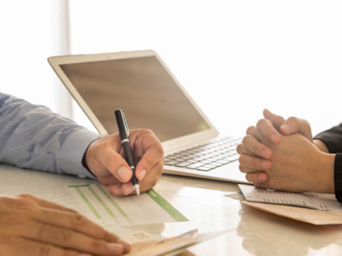 A laptop sits between a financial advisor and a man signing a financial document.