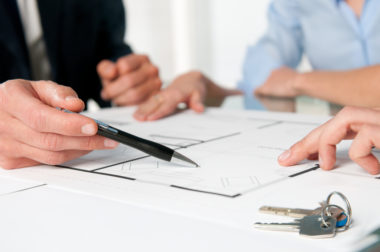 A closeup of a mortgage broker pointing to a document with a pen, showing his client couple a clause in the mortgage.