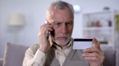 A worried man speaks on the phone while looking at his credit card.