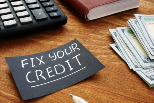A black note sits next to a calculator, notebook, and some cash. The words "fix your credit" are written on in in white ink.